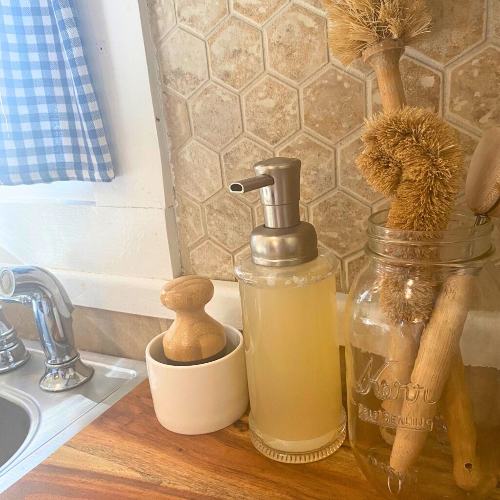 Natural dish soap glass pump jar, small white soap dish with scrub brush, mason jar with scrub brushes next to kitchen sink