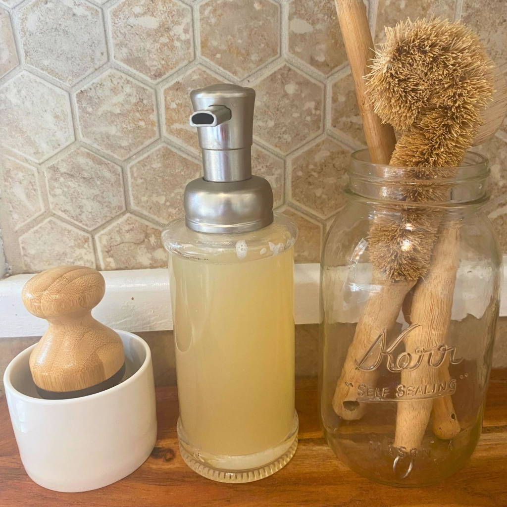 Natural dish soap in glass pump jar, mason jar with scrub brushes, and small white soap dish with brush