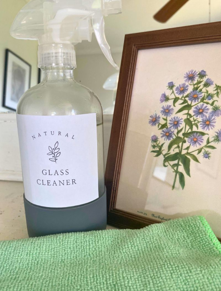 DIY Glass cleaner in clear spray bottle next to mirror, green microfiber cloth, and art