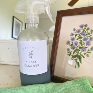 DIY Glass cleaner in clear spray bottle next to mirror, green microfiber cloth, and art